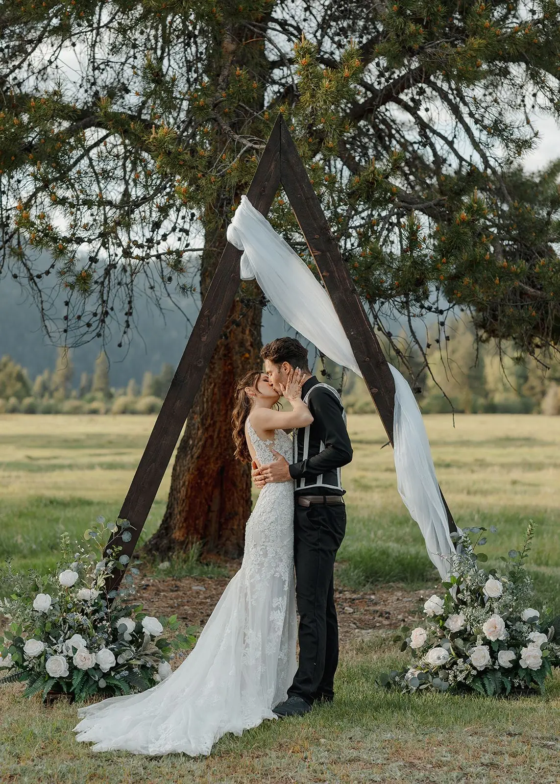 A couple kissing under the ceremony arch, framed by a tall pine