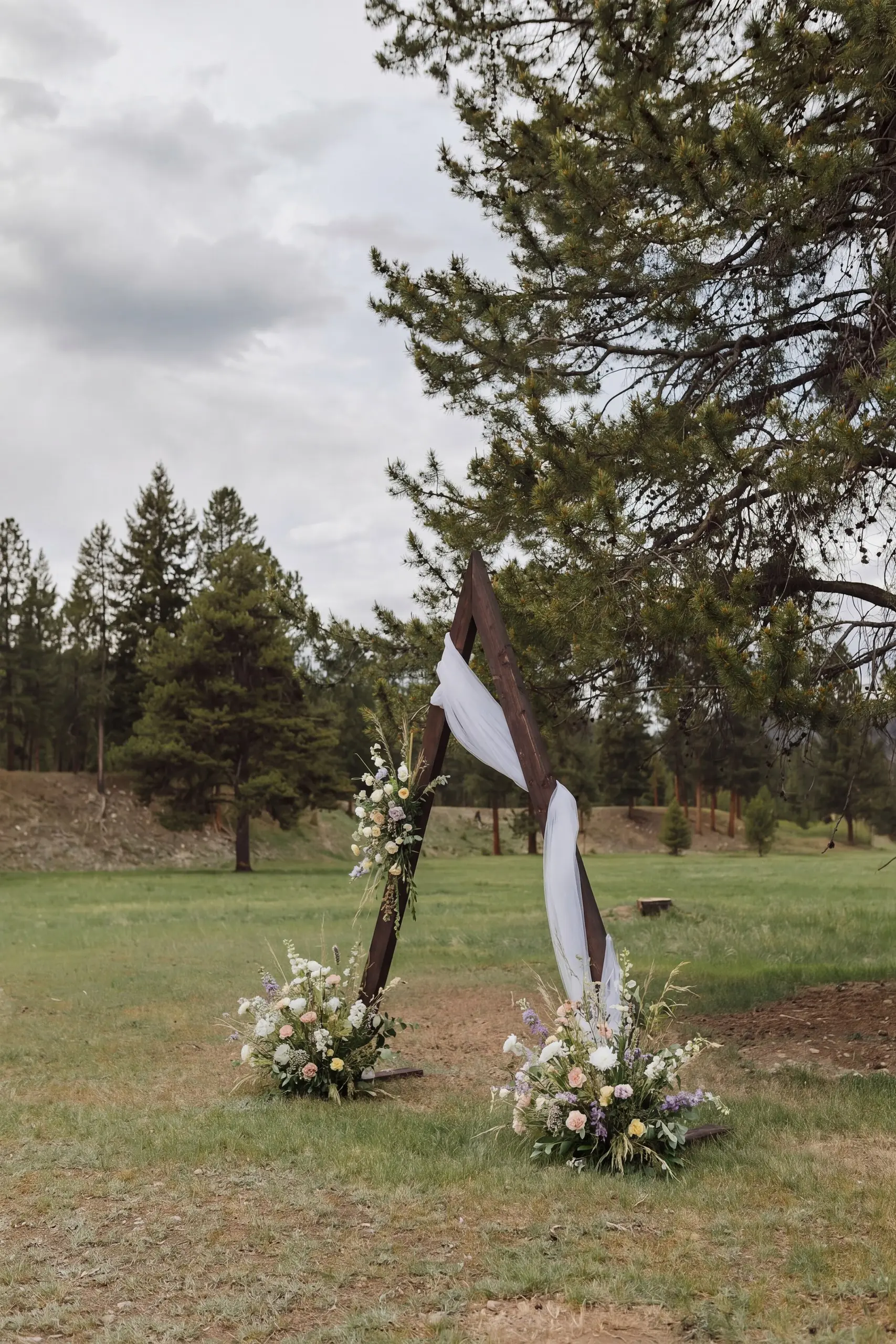 A simple wooden arch with white fabric and wildflowers at the edge of the meadow, pines behind it